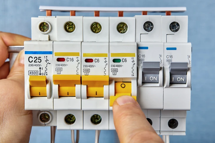 Photographie d'un électricien portant un uniforme professionnel et un badge d'identification, intervenant rapidement à domicile chez un client. Il tient un multimètre ou un autre outil de diagnostic et effectue une intervention électrique (mise aux normes, installation, réparation). Son fourgon de service blanc ou gris portant un marquage professionnel discret est visible en arrière-plan, garé devant la maison du client. La scène transmet l'urgence, la compétence professionnelle et la présence locale - un électricien réactif et qualifié qui arrive rapidement pour résoudre un problème électrique.