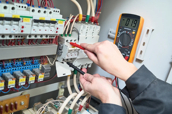 Électricien en intervention sur installation électrique domestique, utilisant un multimètre à pince jaune pour mesurer l'intensité du courant dans un tableau électrique