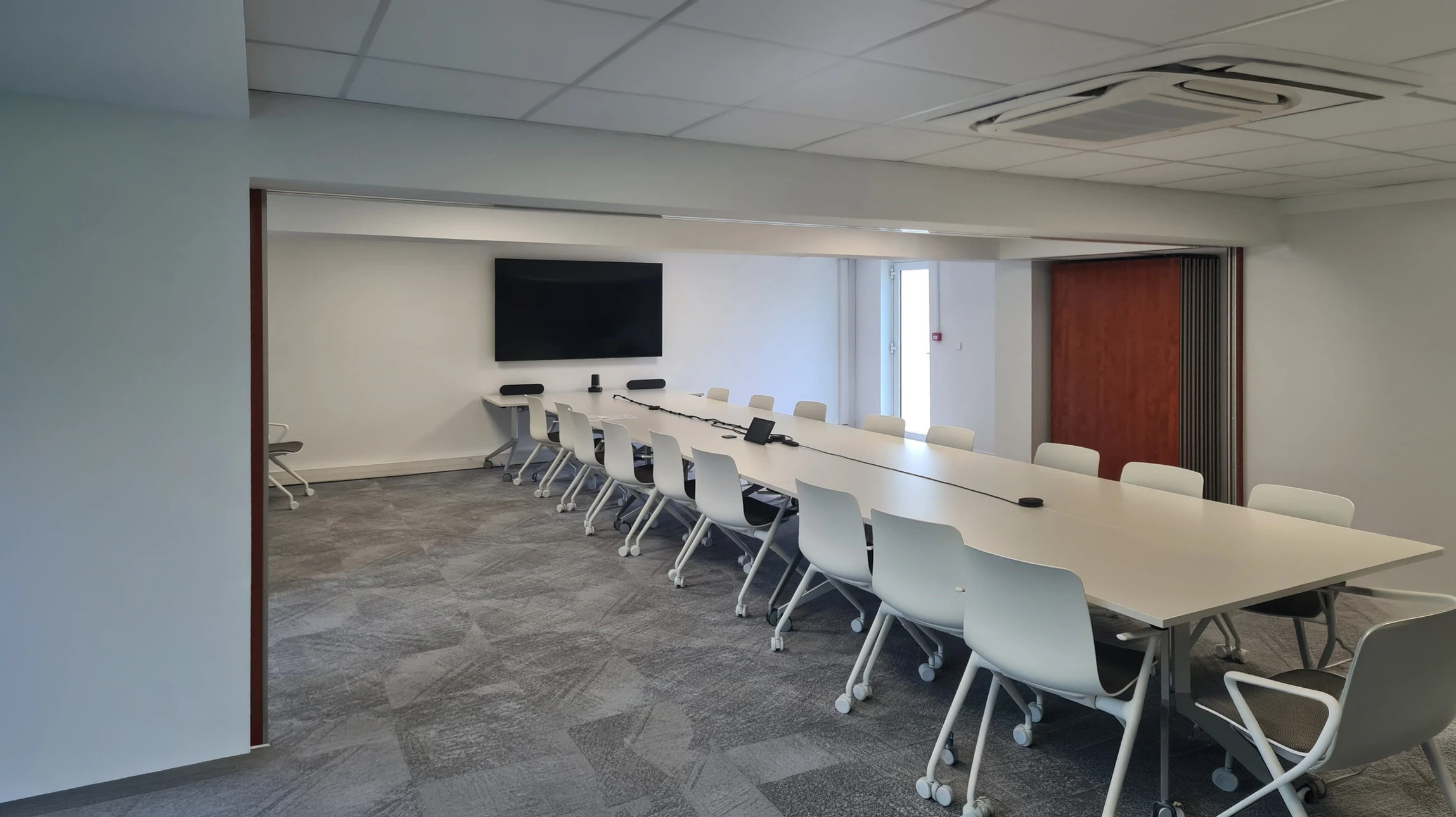 Salle de conférence moderne et élégante vue de face, avec une grande table centrale en bois clair entourée de chaises ergonomiques. Au mur face à la table, un grand écran de projection ou écran plat dernier modèle affichant une présentation professionnelle. Des appareils de vidéoconférence et des haut-parleurs intégrés sont visibles. L'éclairage est professionnel avec des led discrètes. Deux ou trois professionnels en tenue de travail sont positionnés dans la salle, en train d'interagir avec l'équipement ou de discuter. L'ambiance générale est moderne, fonctionnelle et haut de gamme, illustrant une salle connectée pour vidéoconférence et réunions professionnelles.