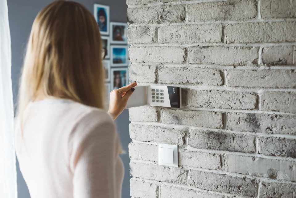 Un homme en chemise à carreaux bleus installe ou configure un panneau de commande d'alarme de sécurité domestique sur un mur blanc intérieur